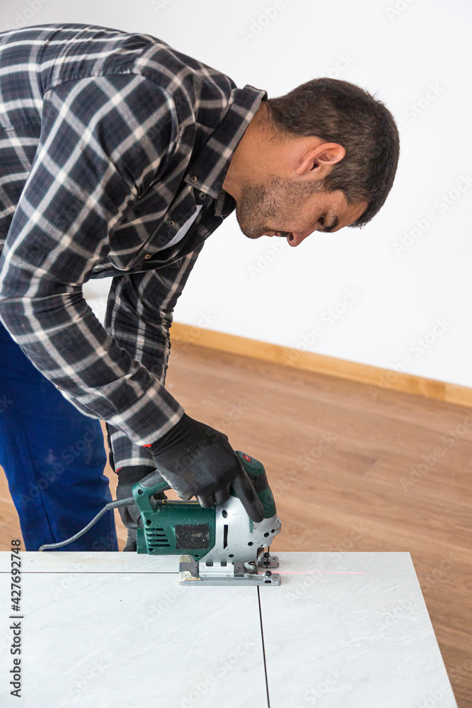 A carpenter with a corded jigsaw cutting a part of the wooden surface (board)
