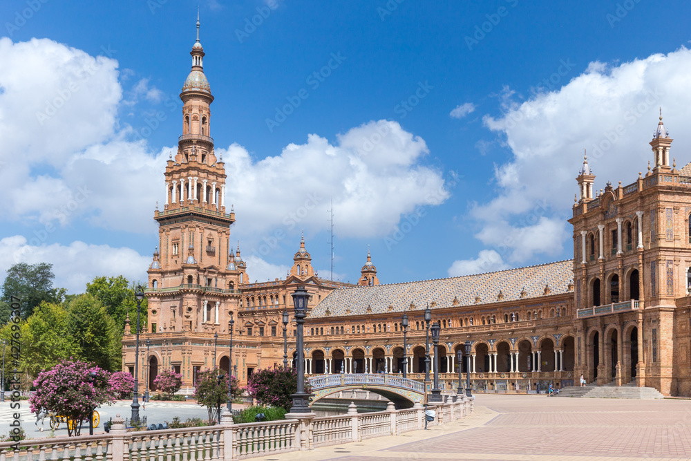 Fototapeta premium Spanish Square, in the center of Seville. Very touristic travel destination empty due to coronavirus measures. View of the south tower. Coloured flowers, blue sky with few clouds. 