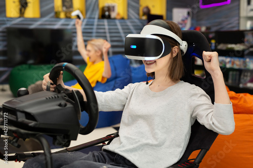 Teenage girl in virtual reality glasses holds the steering wheel and plays a computer game on the console, rejoicing at the victory.
