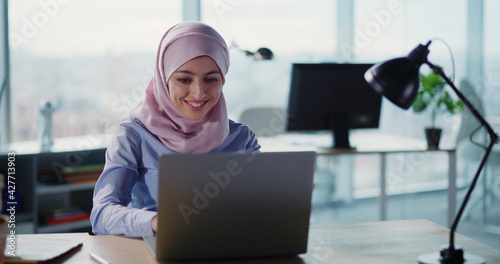 Canvas Print Muslim beautiful young woman in hijab sitting in coworking office workspace typing keybpoard on laptop computer device indoors