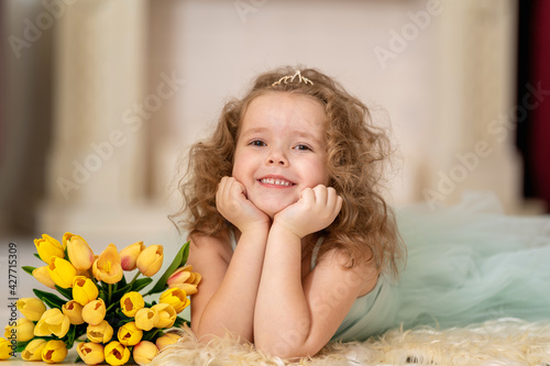 Portrait cute little girl in beautiful mint-colored dress with wavy hair and a tiara. baby is smiling happily and enjoying the flowers in the background of the fireplace, with a bouquet of tulips.