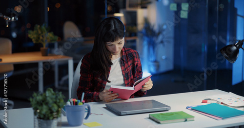 Young caucasian woman with black hair sitting in corporate office reading journal books preparing for worlk. Night routine. Nightlife.