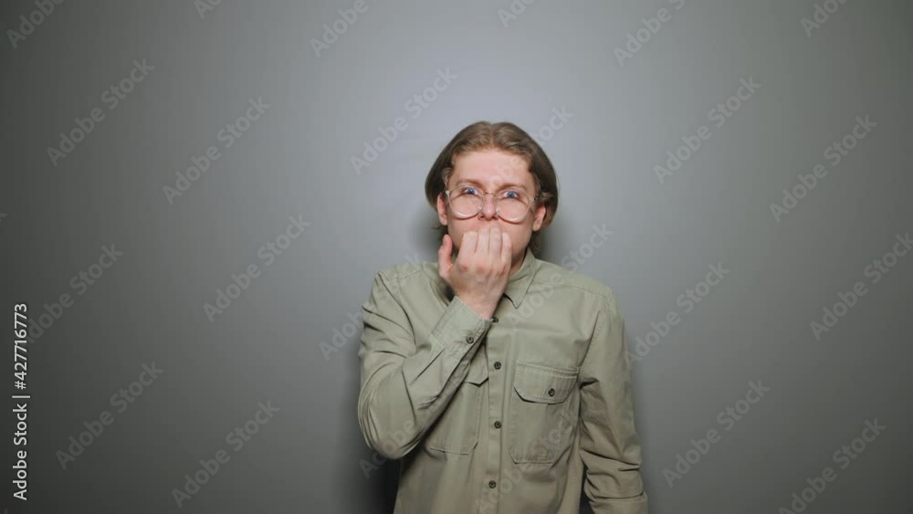 Nervous young man in glasses and shirt bites his nails and looks intently at the camera on a background of a gray wall. Nervous young man in glasses and shirt bites his nails and looks intently at the camera on a background of a gray wall.