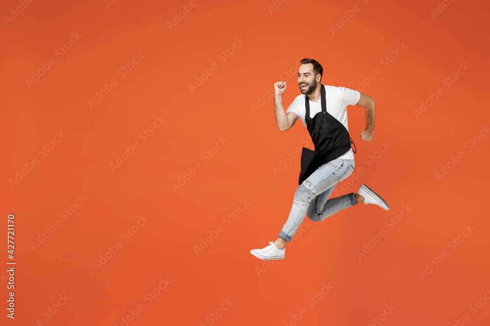 Full length side view young fun man barista bartender barman employee in black apron white t-shirt work in coffee shop jump high run fast isolated on orange background. Small business startup concept.