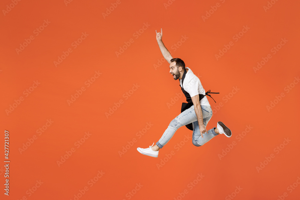 Full length young man barista bartender barman in apron white t-shirt work in coffee shop jump horns up gesture, depicting heavy metal rock sign isolated on orange background. Small business startup.