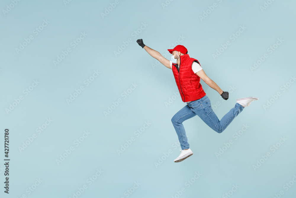 Full length delivery guy employee man in red cap white t-shirt vest uniform sterile face mask gloves work courier service on lockdown covid-19 flu virus run go jump isolated on pastel blue background.