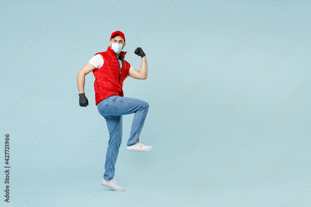 Full length delivery guy employee man in red cap white t-shirt vest uniform sterile face mask gloves work courier service on lockdown covid-19 flu virus run go win isolated on pastel blue background. Full length delivery guy employee man in red cap white t-shirt vest uniform sterile face mask gloves work courier service on lockdown covid-19 flu virus run go win isolated on pastel blue background.