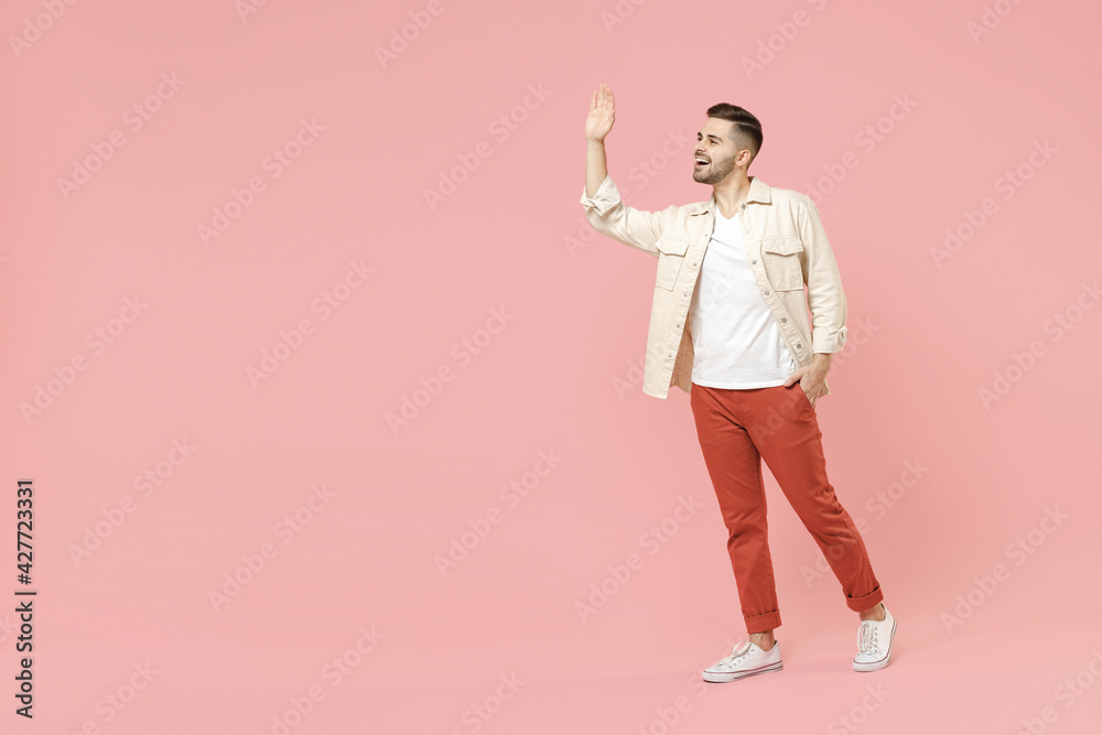 Full length young smiling happy joyful fun trendy fashionable friendly caucasian man 20s wearing jacket white t-shirt walking going waving hand hello isolated on pastel pink color background studio. Full length young smiling happy joyful fun trendy fashionable friendly caucasian man 20s wearing jacket white t-shirt walking going waving hand hello isolated on pastel pink color background studio.