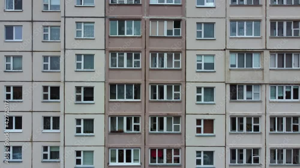 The drone slowly descends near a multi-storey panel house in the style of the USSR. Soviet style architecture. Typical residential building in the ghetto in a post-Soviet city with colored windows. The drone slowly descends near a multi-storey panel house in the style of the USSR. Soviet style architecture. Typical residential building in the ghetto in a post-Soviet city with colored windows.