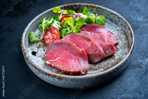 Modern style gourmet raw Japanese tataki beef filet with lettuce and tomatoes as close-up on a Nordic design plate with copy space