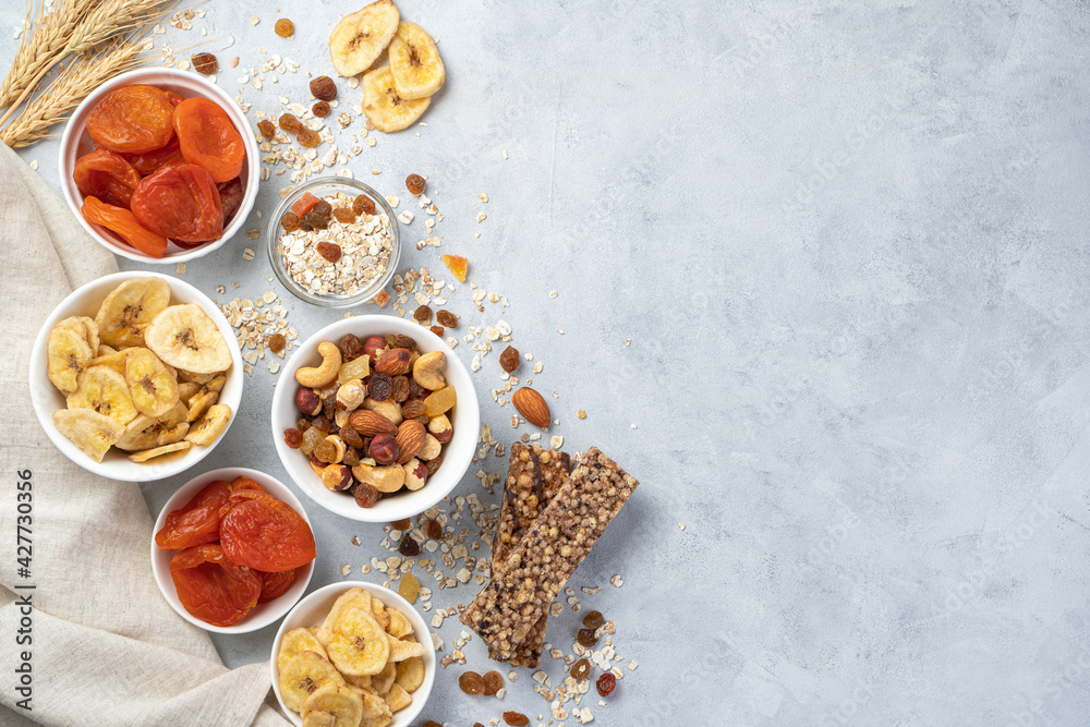 Fototapeta premium Healthy, vitamin-rich, sweet snacks. Banana chips, dried apricots, different types of nuts, raisins and granola bar on a gray background.