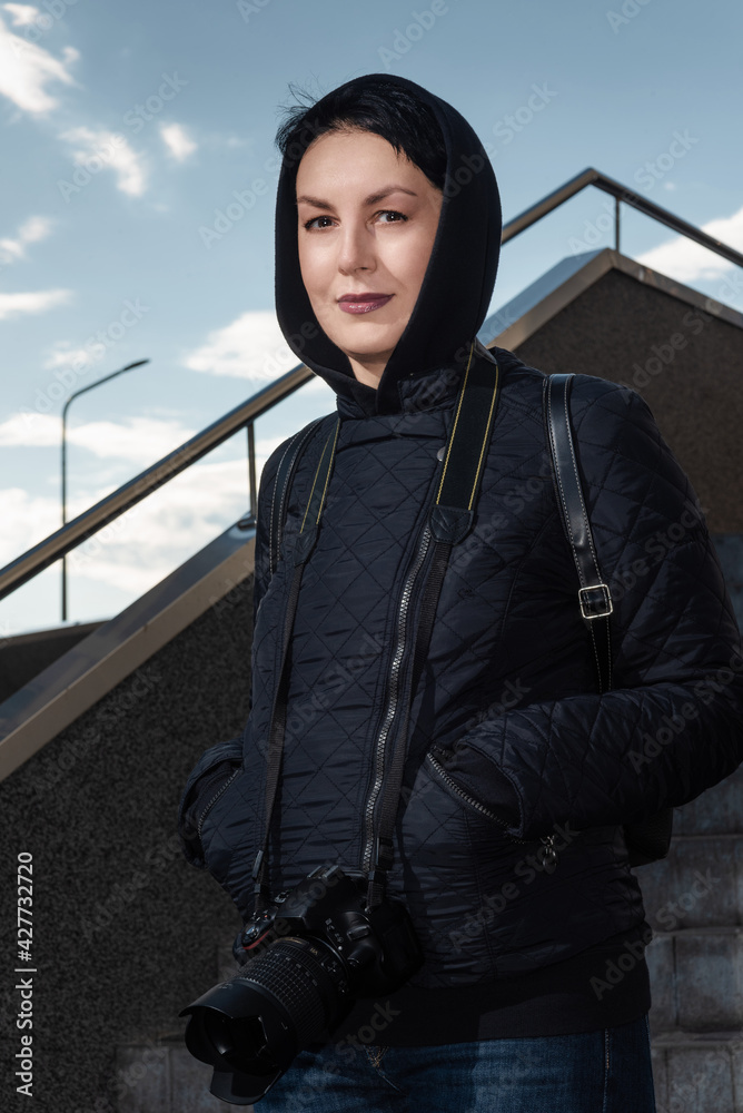 Smiling middle-aged female photographer with professional dslr camera on her neck. Confident woman in hood on the head standing on stairs looking at camera outdoors.