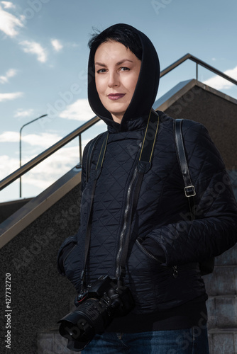 Obraz na płótnie Smiling middle-aged female photographer with professional dslr camera on her neck. Confident woman in hood on the head standing on stairs looking at camera outdoors. w salonie