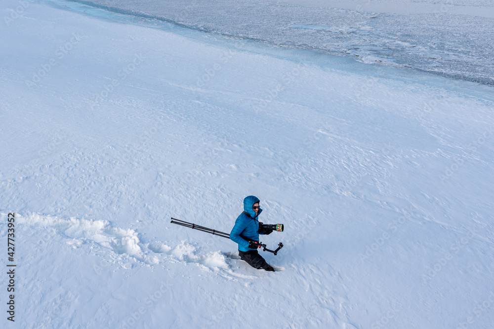 A man, photographer walking across a winter landscape with very deep snow with camera, tripod and a blue jacket with white background. 