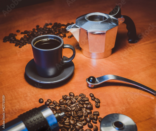 Freshly brewed cup of coffee from a moka pot, set on table in-between coffee grinder, coffee beans ant top part of moka pot.