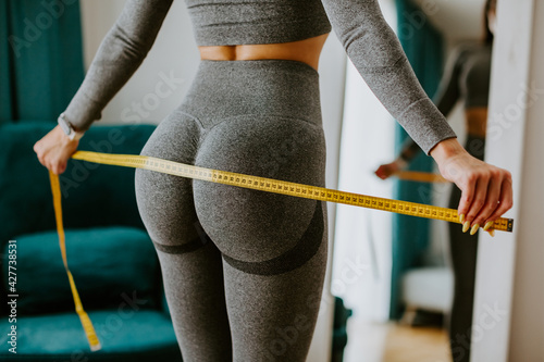 Fitness Woman Measuring body With Tape Standing In Front Of Mirror At Home. Beautiful woman checking success of weight loss program and measuring butt in front of mirror in living room. Slimming
