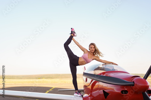 Athletic woman practicing stretching near a red private plane. Working out on the aerodrome on planes and sky background.