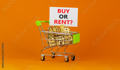 Buy or rent real estate symbol. Miniature shopping cart with wooden houses, words buy or rent. Beautiful orange background, copy space. Business and buy or rent real estate concept.