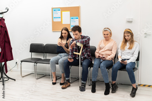 Patients sit in a clinic waiting for their turn to go to the doctor's office. They look at their cell phone while waiting.