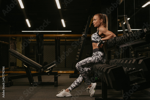 A photo from the side of a fit woman with blonde hair who is sitting on the row of dumbbells in a gym. A girl is relaxing after a workout.