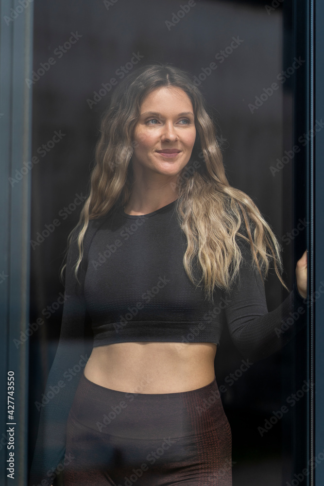Woman in sportswear happy smiling by the window at home. Sporty active attractive fitness woman looking outside Woman in sportswear happy smiling by the window at home. Sporty active attractive fitness woman looking outside