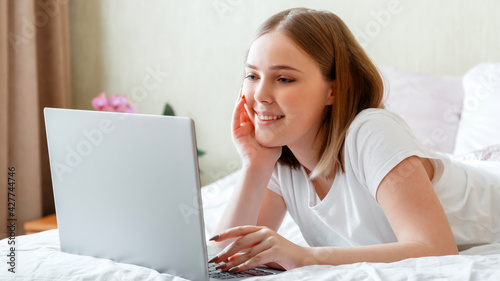 Young woman smile and work using laptop while lying in bed in morning at home. Happy girl in pajamas studying online or planning her day use computer laptop in morning time in bedroom. Long web banner