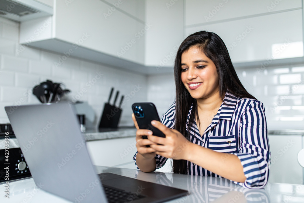 Young indian woman sits at the kitchen table using a laptop and use on a phone. Young indian woman sits at the kitchen table using a laptop and use on a phone.