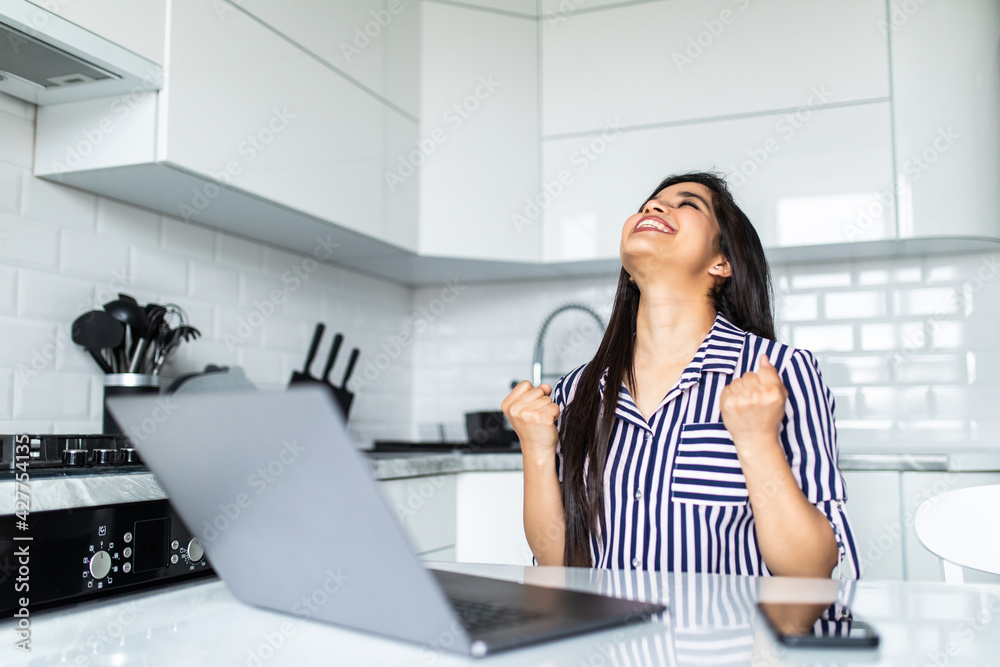 Excited young indian woman with win gesture sitting at table and looking at laptop Excited young indian woman with win gesture sitting at table and looking at laptop
