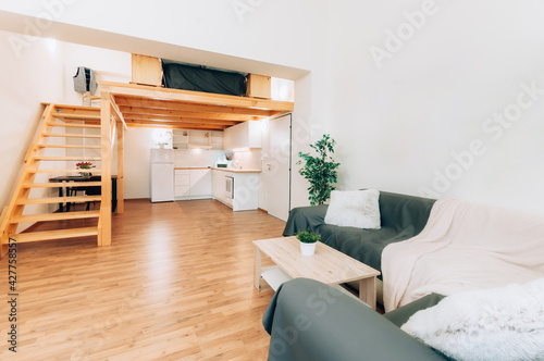 Loft apartment with kitchen, which is under wooden construction forming second floor with staircase. Room features decorative plants, set dining table and regular kitchen appliances.