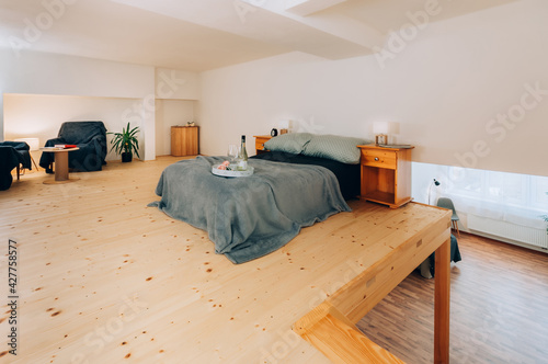 Ordinary bedroom in loft apartment in wooden style with double bed, bedside tables and two glasses of wine with bottle on tray. In corner are two armchairs with small table and decorative plant.