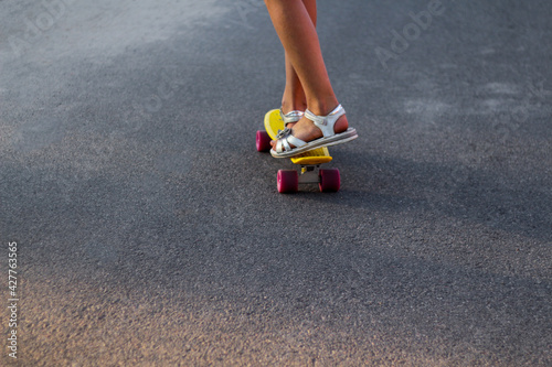 Defocus skater moving along asphalt road on a yellow skateboard wearing white sandals. Some of the legs are on a skateboard close up. Young sport activity. Leisure activity. Penny board. Out of focus