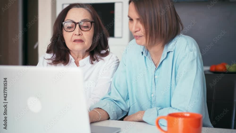 Adult daughter and her mature mother looking at the laptop screen and making online shopping. Smiling young woman teaching her parent how to use a computer. Adult daughter and her mature mother looking at the laptop screen and making online shopping. Smiling young woman teaching her parent how to use a computer.