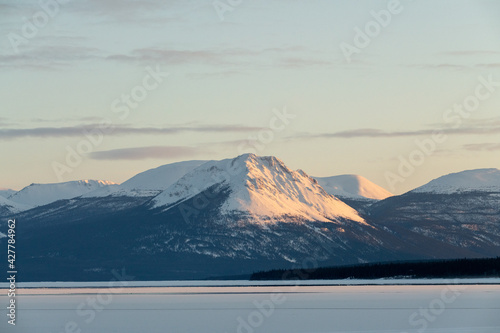 Huge towering snow capped mountains in northern Canada spring time with frozen lake below, wilderness and boreal forest and blue sky background. Taken in Yukon Territory. 