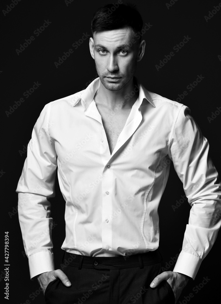 Portrait of pragmatic businessman ceo in official suit trousers and unbuttoned white shirt standing with hands in pockets over black background