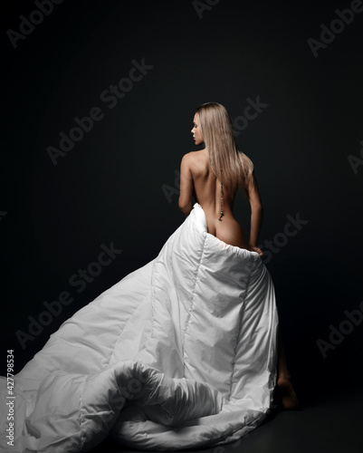 Young sensual sexy naked feminine woman stands back to camera covering herself with large soft white blanket, looking aside over black background. Fashion, vogue, sexy stylish look for woman concept
