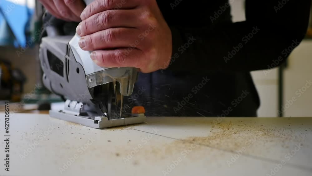 High angle view of skilled carpenter in overalls using speed jig saw to cut small piece of wood. Wood processing. Electric jigsaw cut a wooden board. Cabinetmaker is carving by jig saw a part of wood High angle view of skilled carpenter in overalls using speed jig saw to cut small piece of wood. Wood processing. Electric jigsaw cut a wooden board. Cabinetmaker is carving by jig saw a part of wood