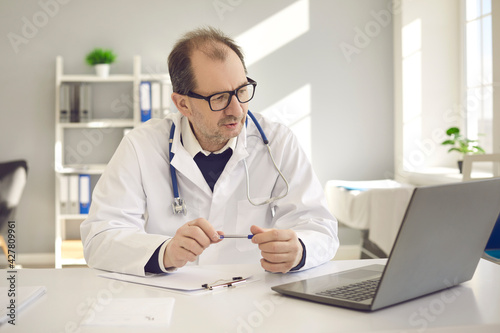 Mature male doctor general practitioner speaking to patient videoconferencing with colleague on laptop having online video call remote consultation at clinic office. Distance medical help telemedicine
