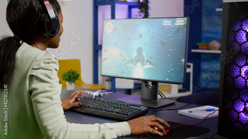 Pro black woman gamer playing space shooter videogame on RGB powerful computer wearing professional headphones. Videogamer streaming gameplay cyber game sitting on gaming chair