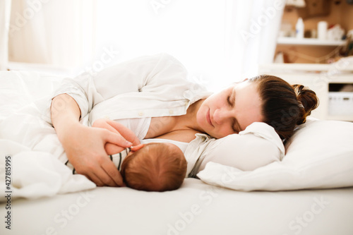 Cute caucasian mom and newborn baby, mom breastfeeds her baby lying on a white background on the bed in the bedroom, close-up, soft focus
