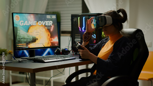 Sad woman gamer wearing virtual reality headset losing space shooter games on online tournement using wireless controller. Defeated player playing for online competition late night in gaming room photo