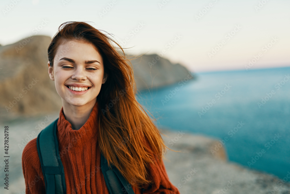 happy woman in a sweater with a backpack on her back smiling on nature in the mountains near the sea