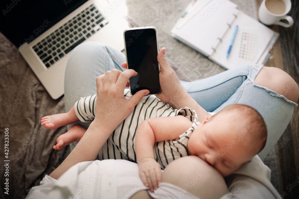 Fototapeta premium Cute caucasian mom and newborn baby, mom breastfeeds her baby and works with phone and laptop. Top view.