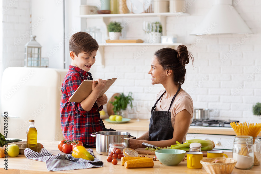 © deniskomarov - Happily smiling mother and son preparing dinner on loft kitchen. Boy sitting on table reading recipe from cookbook to cheerful mom wearing apron. Happy family weekend. Excited housework