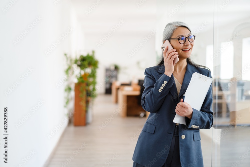 Fototapeta premium The happy Asian woman talking on the phone while looking to the side in a light office