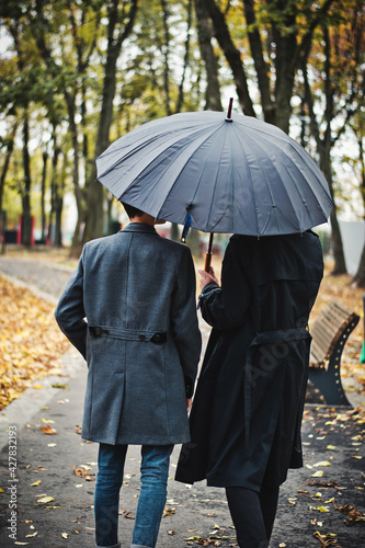 Back view of young gay couple walking in the park in autumn or fall under umbrella