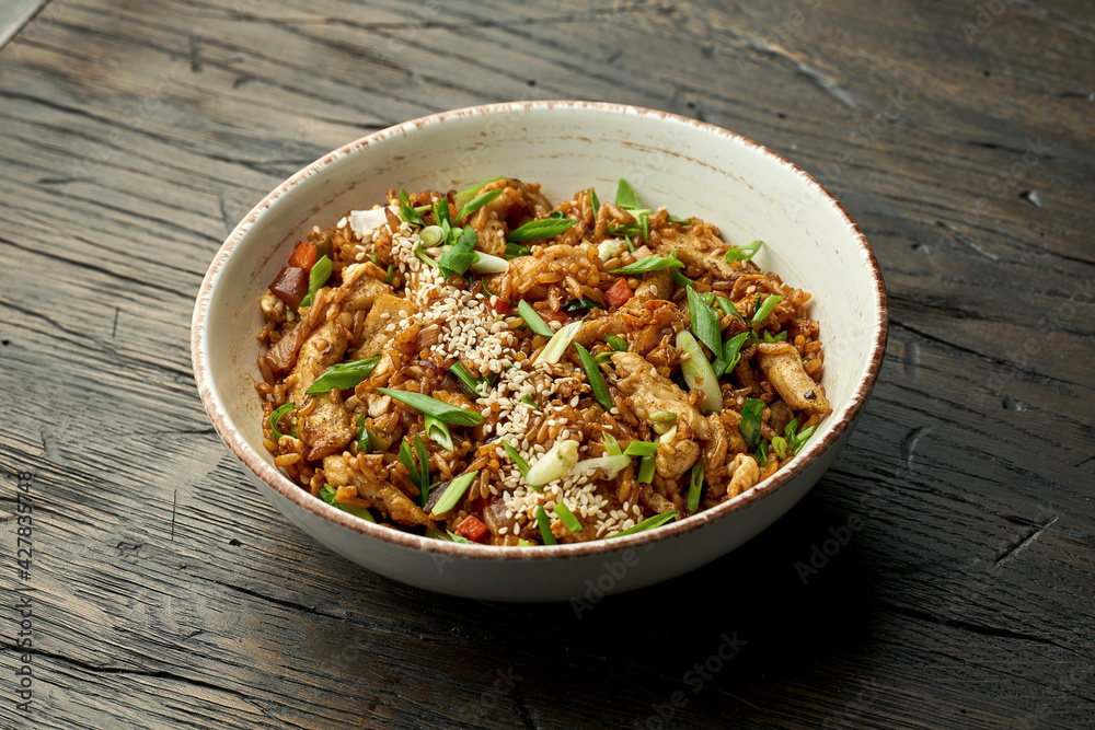 Delicious Asian street food - wok rice with chicken, green onions, vegetables and sesame seeds in a white bowl on a wooden background. Delicious Asian street food - wok rice with chicken, green onions, vegetables and sesame seeds in a white bowl on a wooden background.