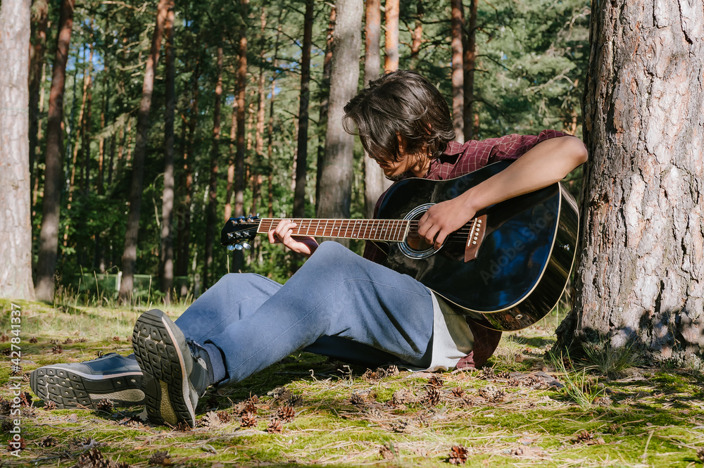 A man plays the guitar while sitting in the forest near a tree. Against the backdrop of nature. A man plays the guitar while sitting in the forest near a tree. Against the backdrop of nature.