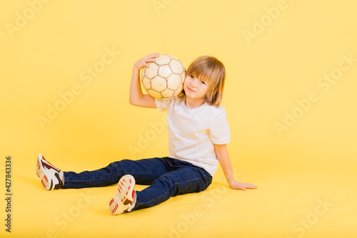 Cute boy is holding a football ball made of genuine leather isolated on a yellow background. Soccer