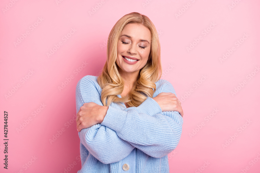 Photo portrait of woman with wavy blonde hair dreamy embracing herself in blue cardigan smiling isolated on pastel pink color background Photo portrait of woman with wavy blonde hair dreamy embracing herself in blue cardigan smiling isolated on pastel pink color background
