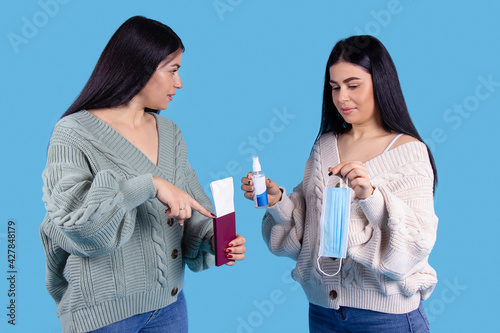 Two twin sisters on a blue background, holding a passport with an airplane ticket, disinfectant and protective masck, informed with regulations during the pandemic. Protection concept. photo